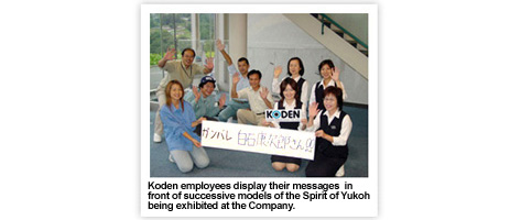 Koden employees display their messages in front of successive models of the Spirit of Yukoh being exhibited at the Company.